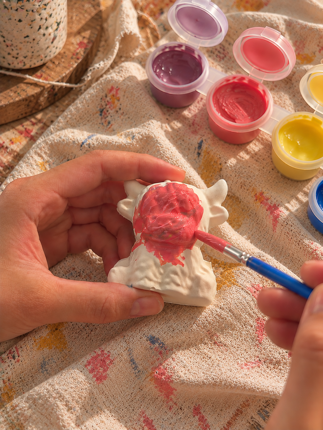 A plaster figurine being painted pink at the craft table, with colourful paint pots nearby